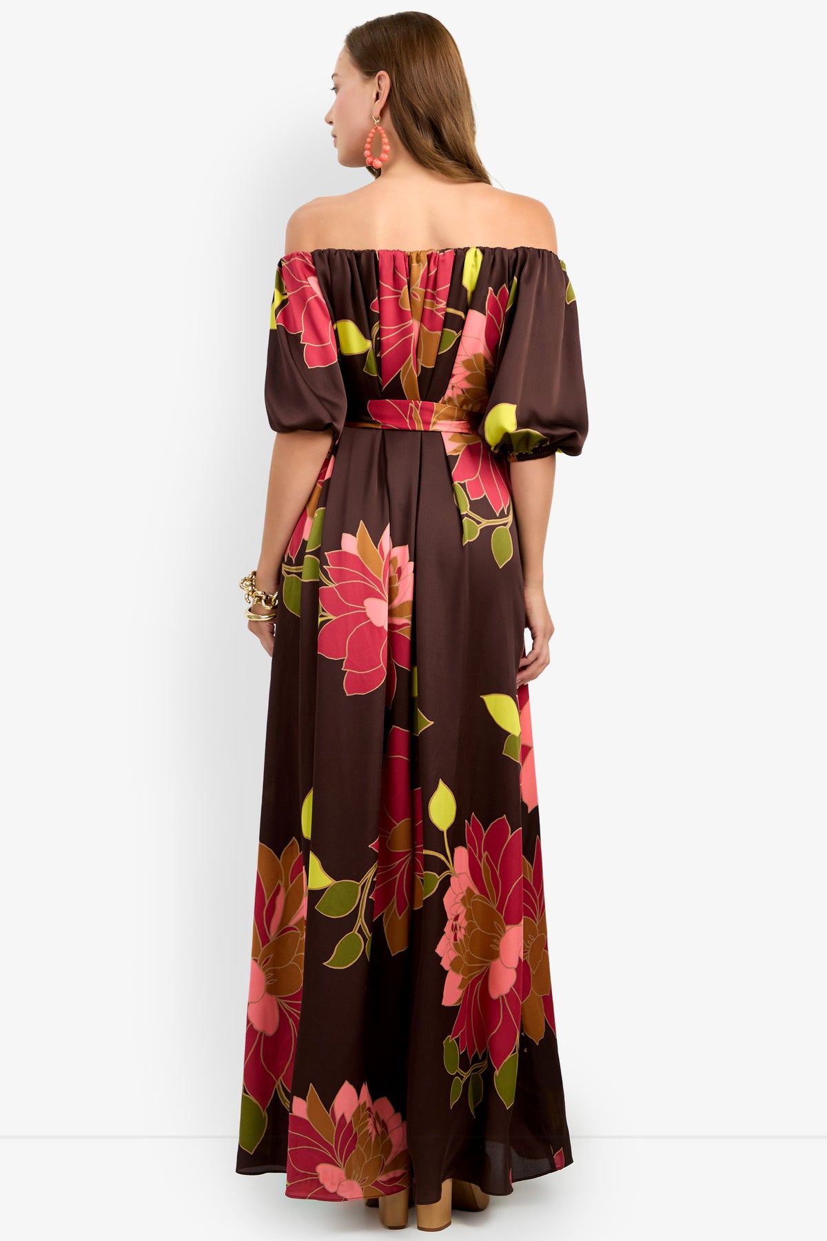 A woman facing away is wearing the Voglia Dress, a brown off-the-shoulder maxi dress featuring large pink, red, and yellow floral patterns. She has long hair, orange earrings, and a bracelet. The background is plain white.