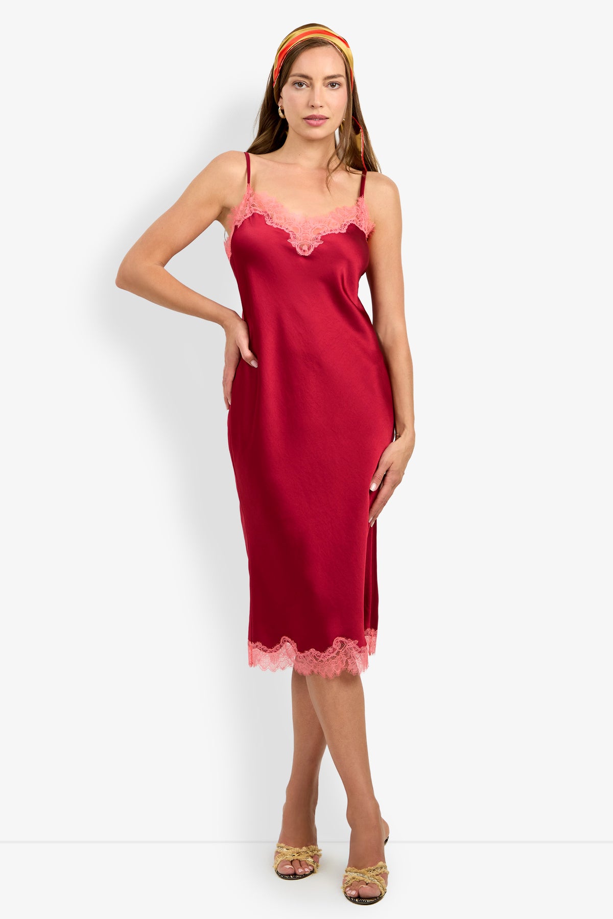 A woman stands against a plain white background wearing the CARAMIA DRESS—a red satin slip dress with pink lace trim, crafted from 100% polyester—paired with a multicolored headband and strappy heeled sandals.