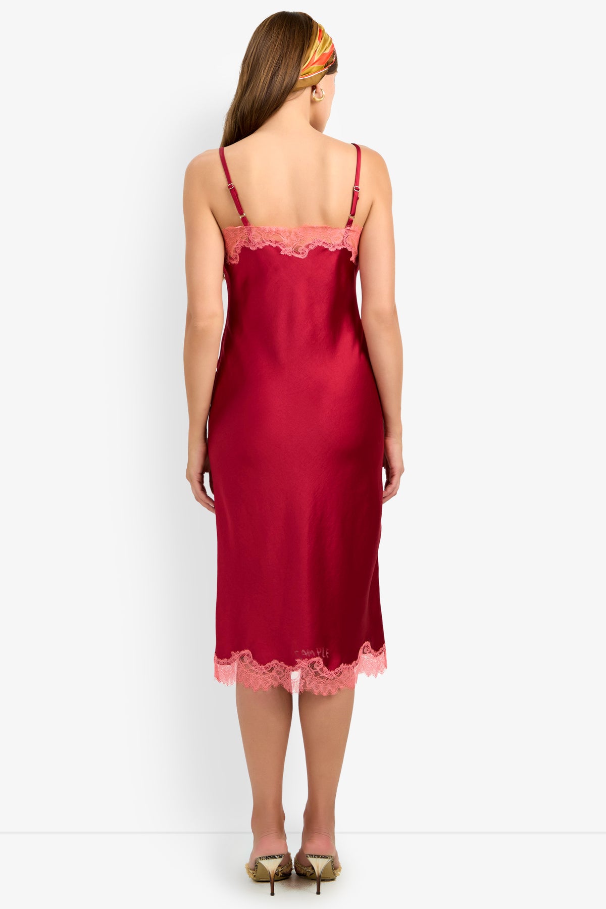 A woman faces away, modeling the CARAMIA DRESS—a red satin slip dress with pink lace trim and adjustable straps, crafted from 100% polyester for a smooth, luxurious feel. She has long brown hair styled with a colorful headband and completes the look with beige heels.
