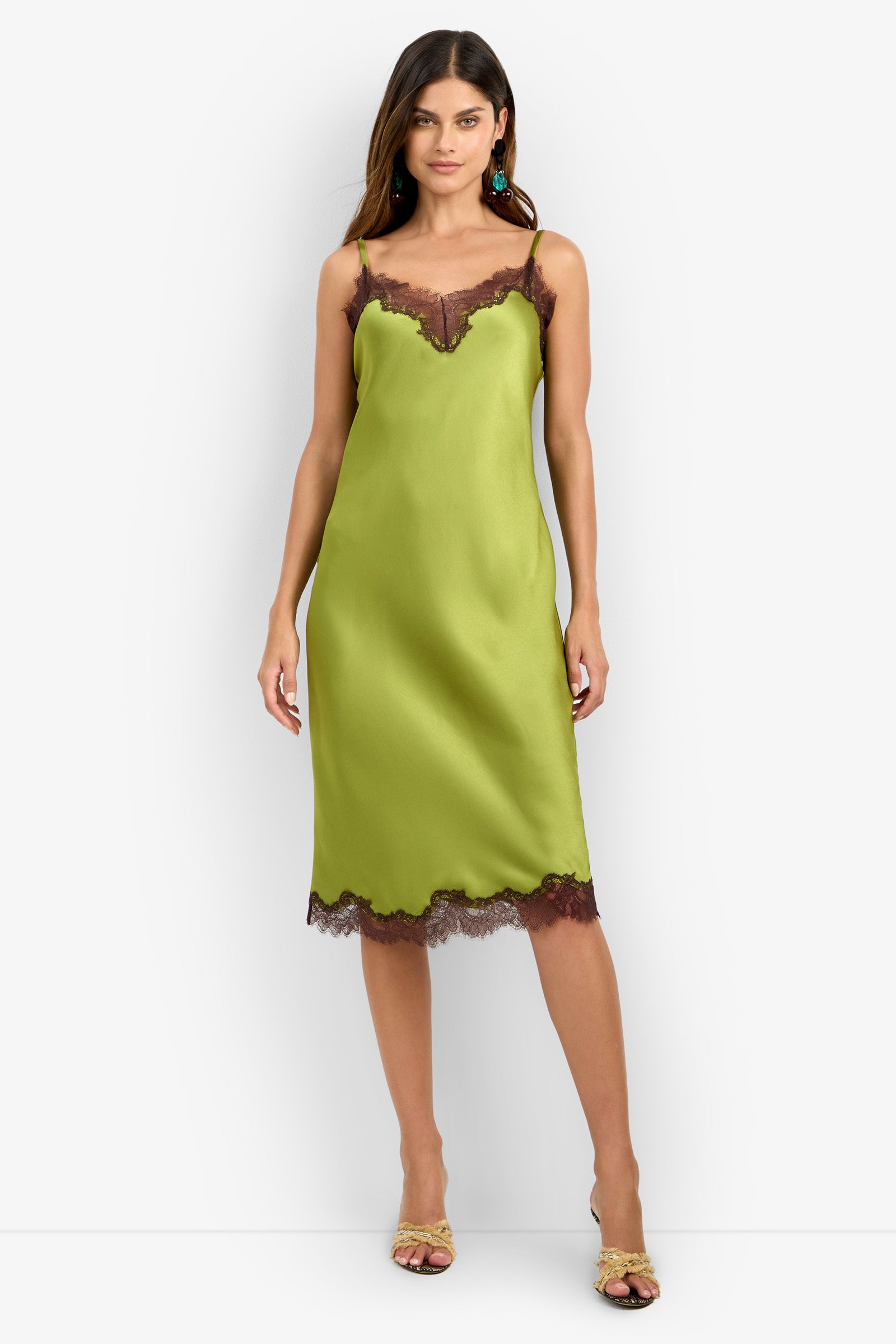 A woman wearing the CARAMIA DRESS, a light green satin slip crafted from 100% polyester with dark lace trim at the neckline and hem, paired with yellow sandals and statement earrings, stands against a white background.