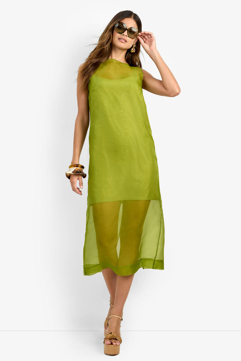 A woman poses against a plain white background with one hand on her head, wearing the ROSELLE DRESS—an airy, sleeveless 100% silk dress in sheer lime green—styled with sunglasses and platform heels.