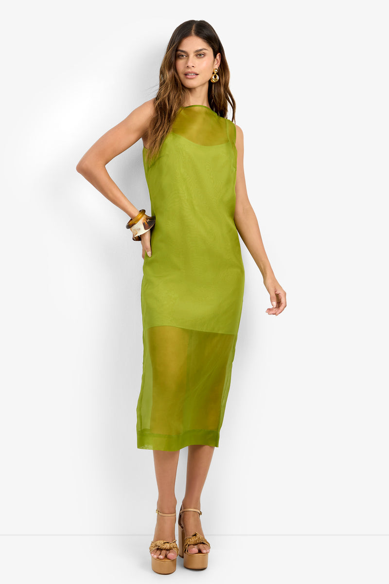 A woman stands against a plain white background wearing the ROSELLE DRESS, a sleeveless, sheer lime green midi dress crafted from 100% silk, paired with tan platform heels and chunky bracelets.