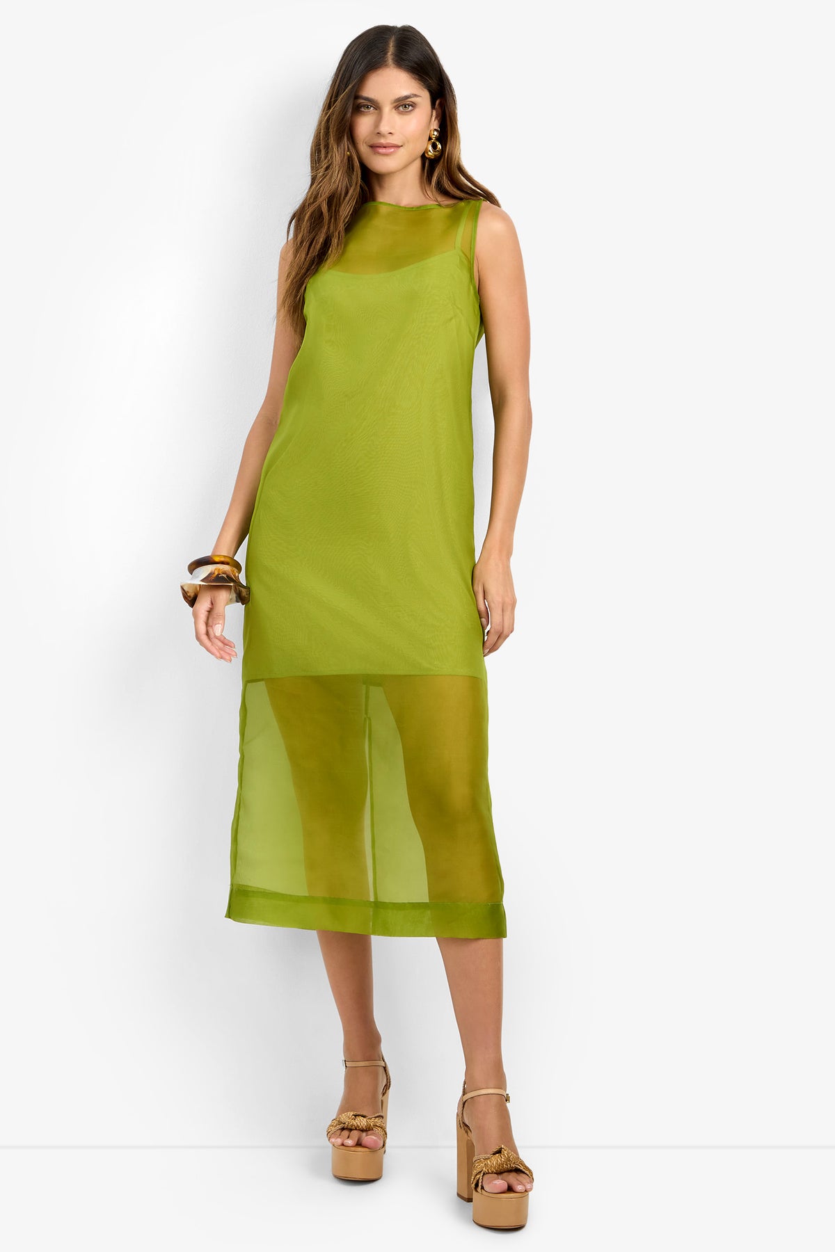 A woman stands against a white background wearing the ROSELLE DRESS—a sleeveless, lime green 100% silk midi dress featuring a sheer lower panel—paired with beige platform heels and chunky bracelets.