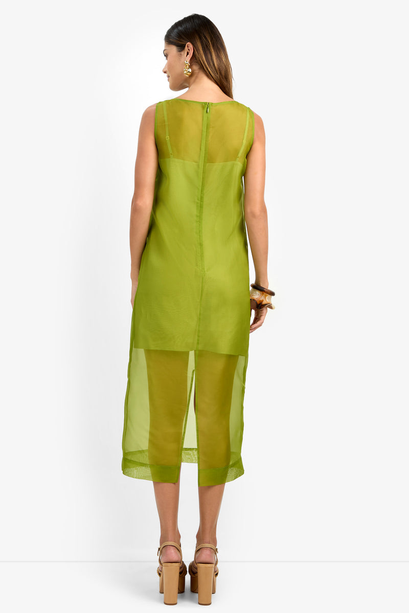 A woman is photographed from the back wearing the ROSELLE DRESS—a sleeveless, sheer green dress crafted from 100% silk—paired with matching pants. She has wavy brown hair, accessorizes with earrings and bracelets, and stands in beige high-heeled sandals against a plain white background.