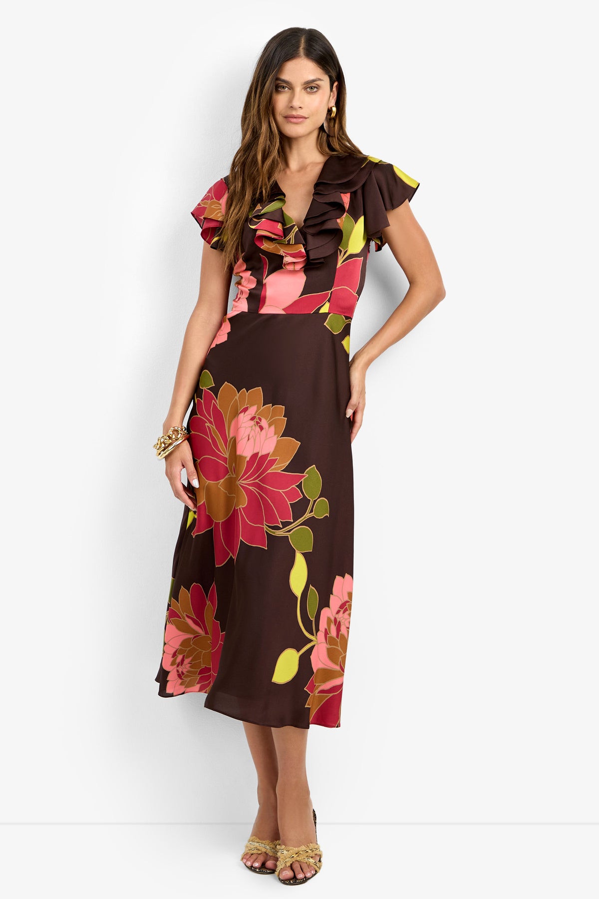 A woman models the PATRONS DRESS, featuring a brown midi length with bold pink and red floral prints, a ruffled neckline, and short flutter sleeves. She has long brown hair and is shown wearing open-toed heels against a plain white background.