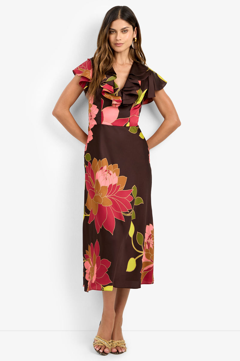 A woman wearing the PATRONS DRESS, a brown midi dress featuring large pink and orange floral prints, a ruffled V-neckline, and short flutter sleeves, paired with tan heeled sandals, stands against a plain white background.
