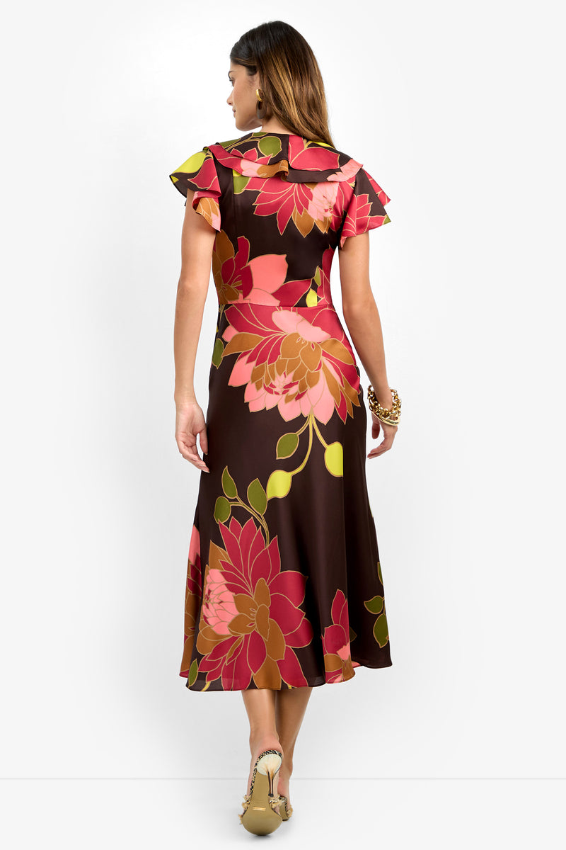 A woman is seen from behind, standing against a white background, wearing the PATRONS DRESS—an ankle-length brown dress adorned with large pink and red floral patterns and featuring short flutter sleeves. She completes the look with beige heels.