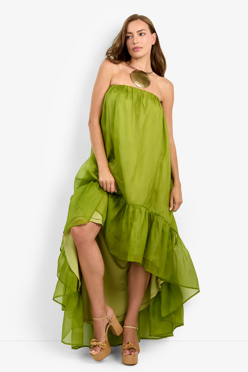 A woman models the ARIAH DRESS, a strapless lime-green high-low ruffle dress, paired with tan platform heels and a bold round gold statement necklace against a white background.