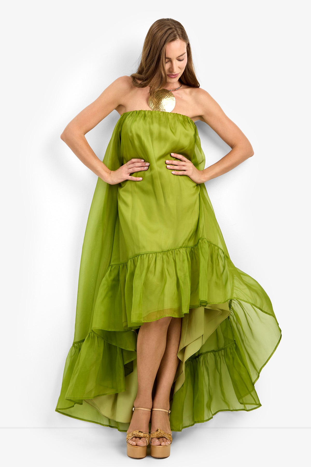 A woman wears the ARIAH DRESS—a strapless, high-low lime green ruffled design—and tan platform heels as she stands against a plain white background, looking down and holding the sides of her dress.