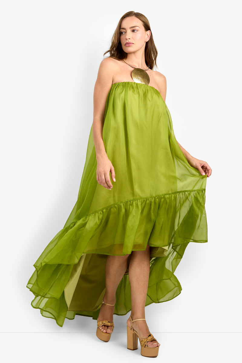 A woman wearing the ARIAH DRESS—a strapless, flowing lime green high-low dress with ruffle detail—pairs it with tan platform heels and a large gold statement necklace as she stands against a plain white background.