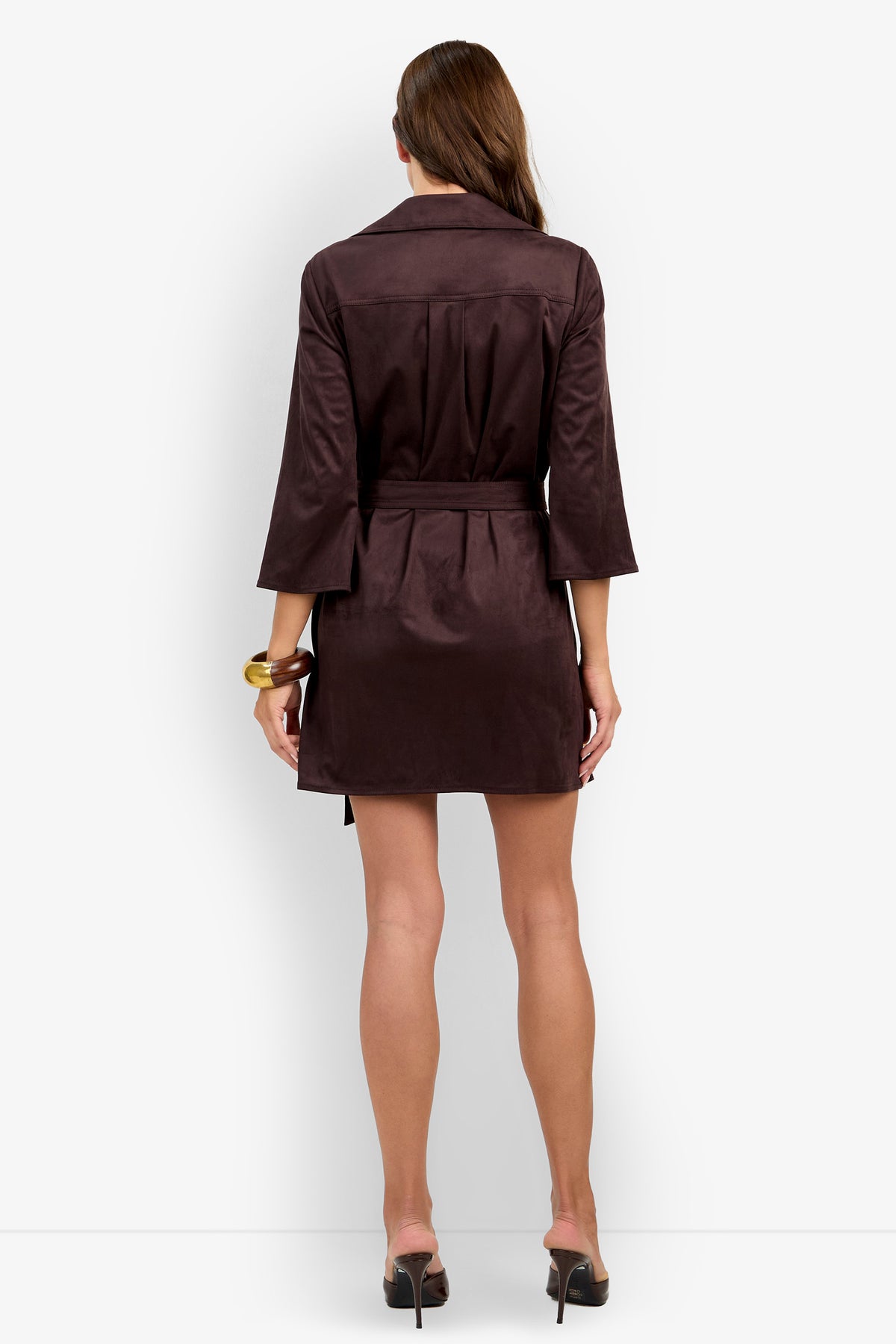 A woman stands facing away, wearing the Annecy Dress—a dark brown belted coat dress with three-quarter sleeves and black heeled shoes—holding a gold bracelet in her left hand.