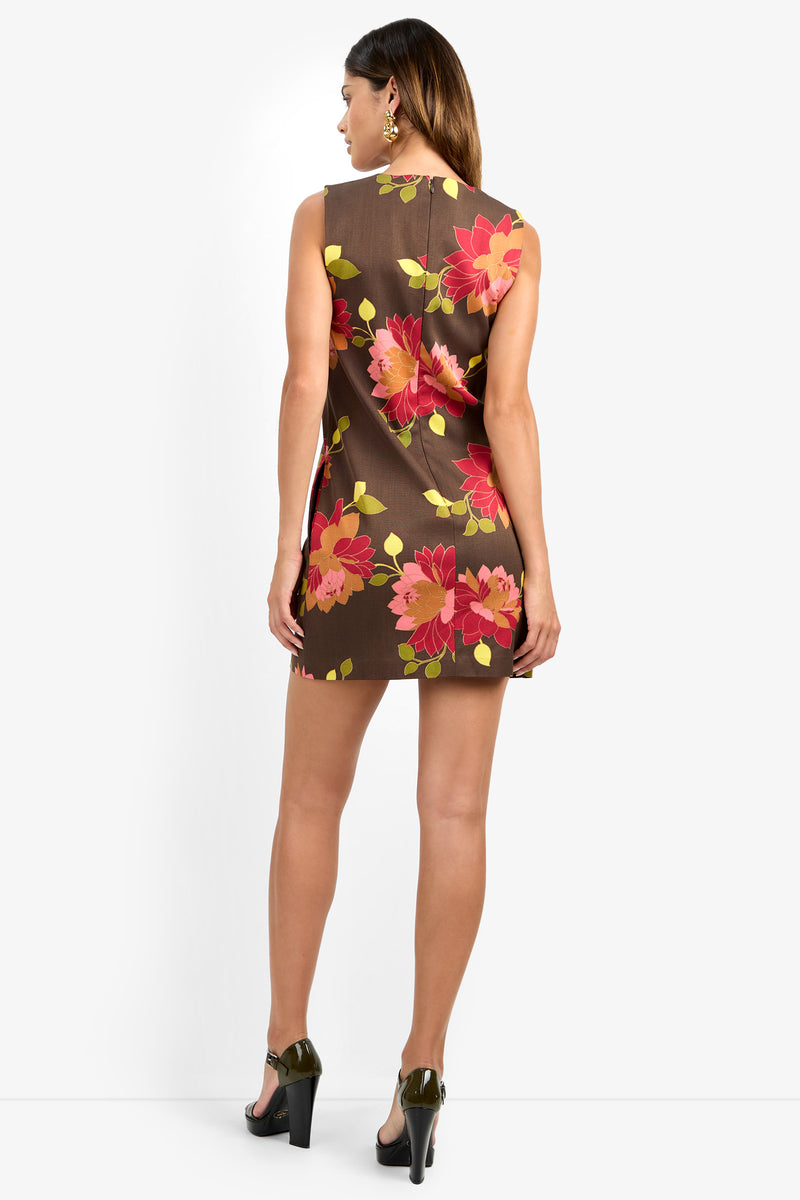 A woman faces away from the camera, wearing the TAHLIA 2 DRESS—a sleeveless brown mini dress featuring bold pink and yellow floral patterns. She has straight brown hair, dark high heels, and earrings, standing against a plain white background.