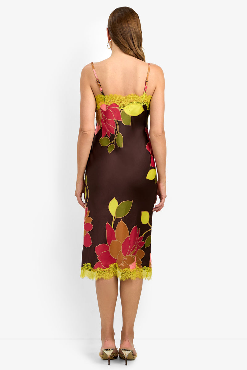 A woman with straight, long brown hair is seen from the back wearing the CARAMIA DRESS (1), a brown midi slip dress featuring bright red, yellow, and green floral patterns, yellow lace trim, and thin straps. She pairs the look with beige heeled sandals.