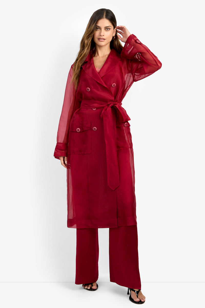 A woman stands against a plain white background wearing the ANGELICO TRENCH, a sheer red trench coat with a tie belt, paired with matching red pants and black heels.