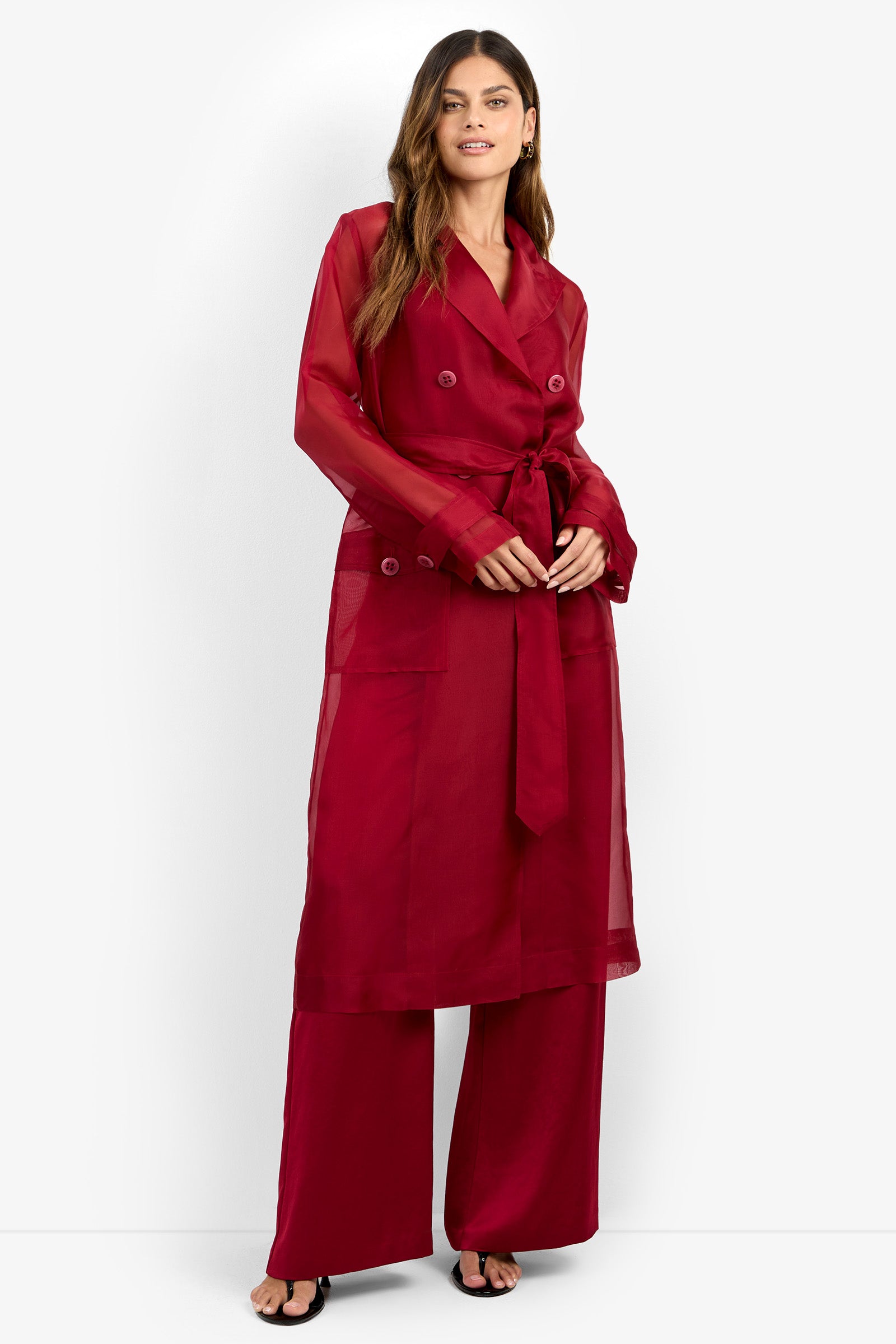 Wearing the ANGELICO TRENCH, a sheer, long red belted coat, over matching wide-leg red pants and black sandals, a woman stands against a plain white background.