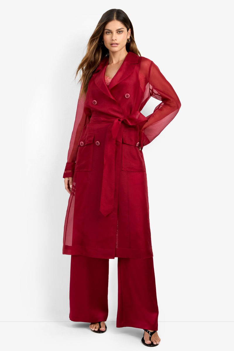 A woman wearing the ANGELICO TRENCH—a sheer, long red trench coat with a belt—paired with matching red pants and black sandals stands against a white background.