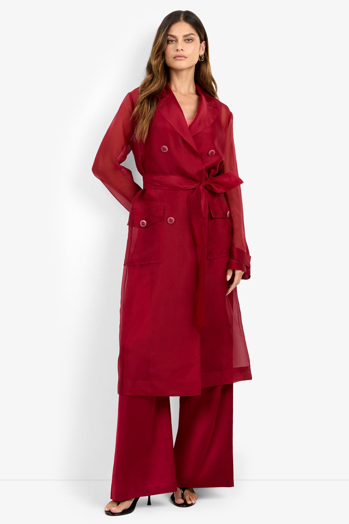 A woman stands against a plain white background wearing the ANGELICO TRENCH, a sheer, long red trench coat with a tie belt, paired with matching wide-leg red pants and black heels.