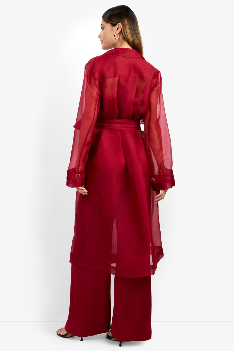A woman is shown from the back against a plain white background, wearing the ANGELICO TRENCH—a sheer, long-sleeve red trench coat—paired with matching wide-leg pants and black heels.