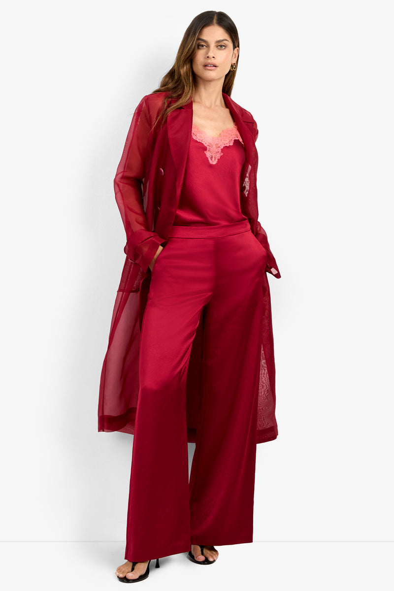 A woman stands against a white background, confidently looking at the camera with her hands in her pockets. She is wearing the ANGELICO TRENCH, a red sheer trench coat, layered over a red satin camisole with lace trim and matching wide-leg pants, paired with black high-heeled sandals.