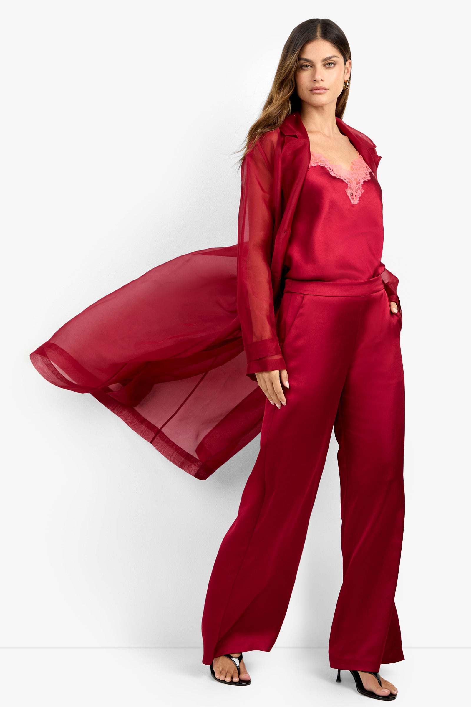 A woman stands confidently against a white background, wearing the ANGELICO TRENCH—a flowing sheer red long coat—over a satin red camisole with lace trim, wide-leg satin red pants, and black heeled sandals.