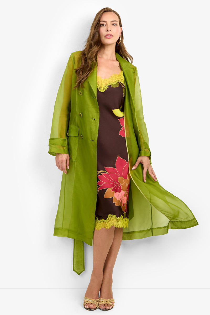 A woman wears the ANGELICO TRENCH, a sheer green trench coat, layered over a brown slip dress with large floral prints and yellow lace, standing against a white background.