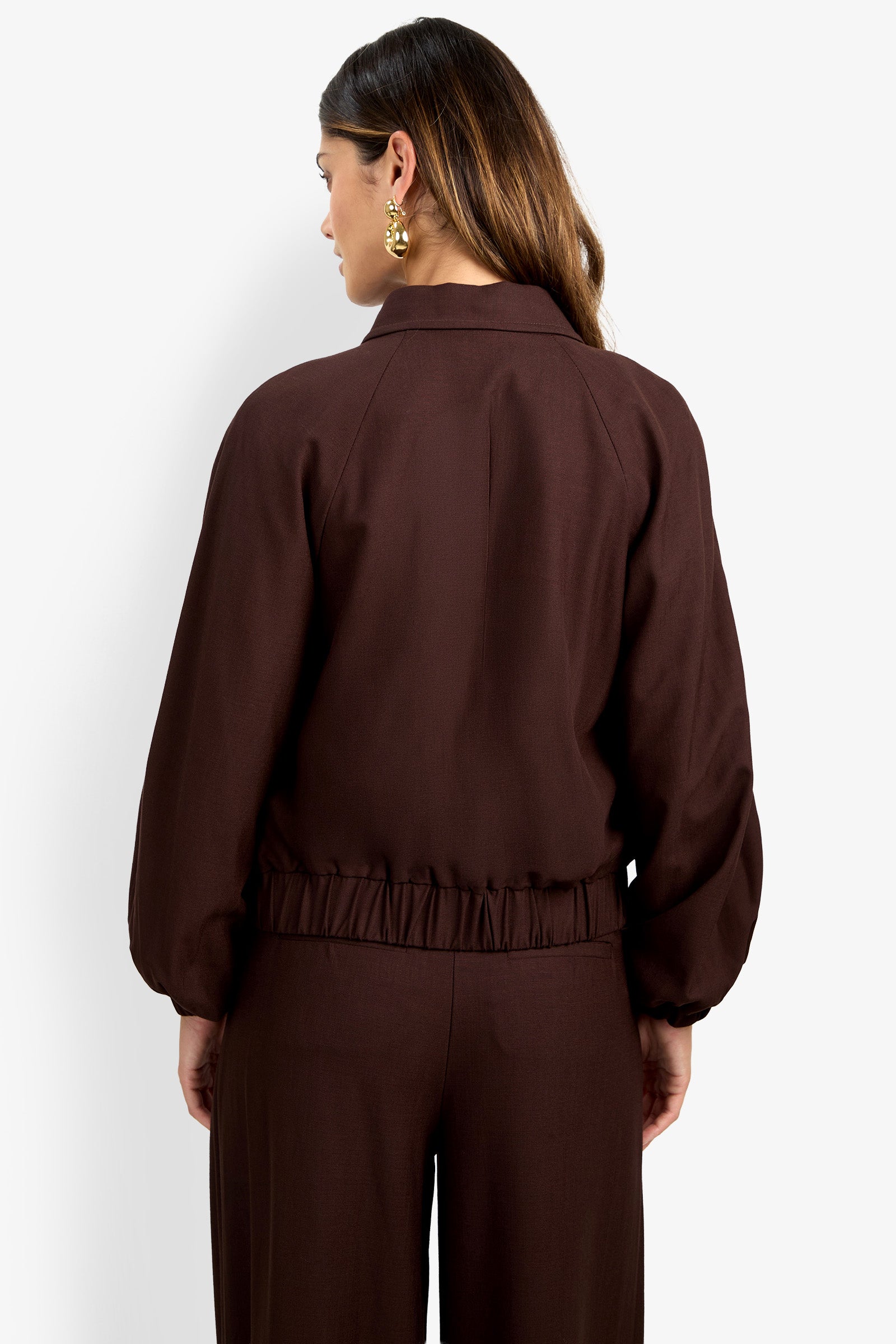 A woman with long brown hair is seen from the back wearing the INFLUENTIAL JACKET—a dark brown, loose-fitting piece with a gathered waist—paired with matching pants and gold earrings.