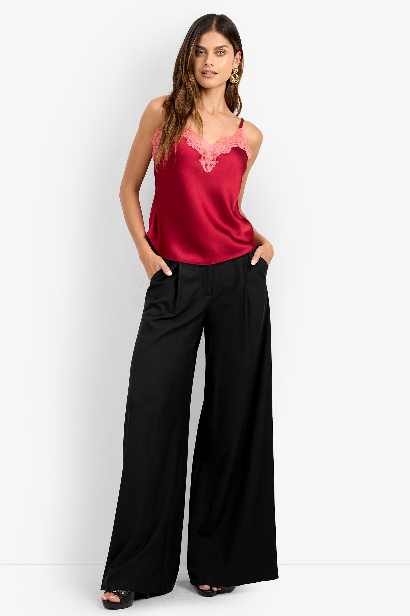 A woman with long wavy brown hair and gold hoop earrings wears a red lace-trimmed camisole, platform sandals, and high-waisted, wide-leg black MIMMO PANT with her hands in her pockets.