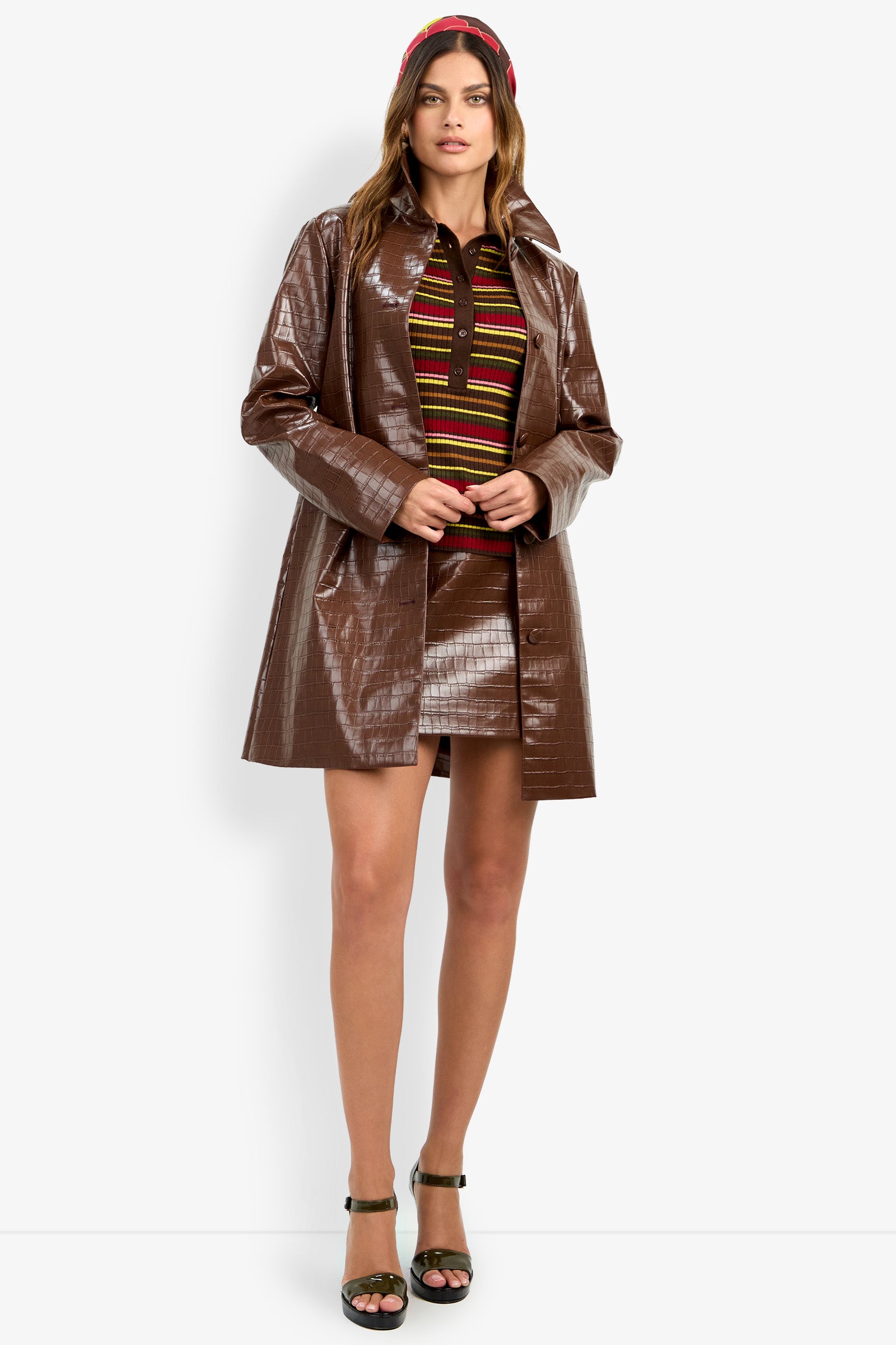 A woman stands against a white background wearing the DIVIAN COAT—a shiny brown coat—paired with a skirt set, striped top, red headband, and dark green high-heeled sandals.