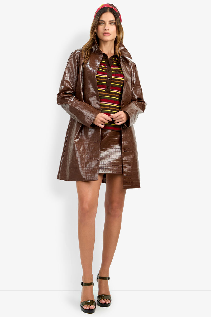 A woman stands against a white background wearing the DIVIAN COAT—a shiny brown coat—paired with a skirt set, striped top, red headband, and dark green high-heeled sandals.