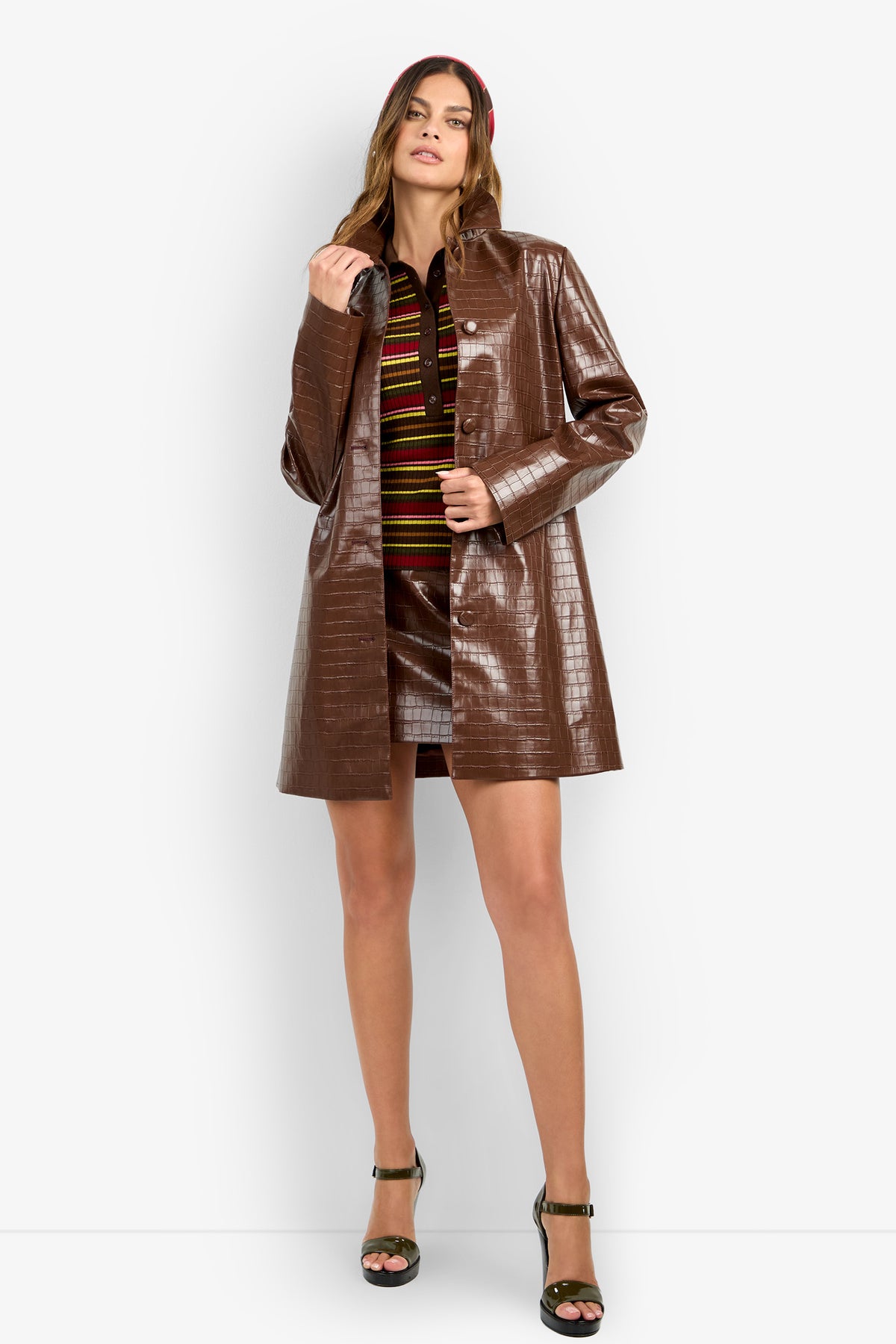 WOMAN standing and wearing the DIVIAN COAT—a shiny brown textured piece—over a striped shirt and matching brown mini skirt, paired with green high-heeled sandals. She poses with one hand holding the coat's lapel.