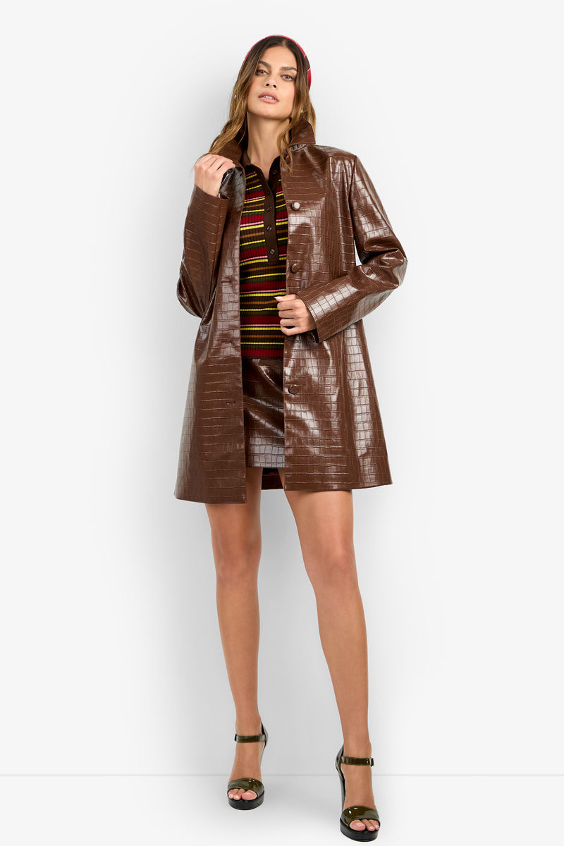 WOMAN standing and wearing the DIVIAN COAT—a shiny brown textured piece—over a striped shirt and matching brown mini skirt, paired with green high-heeled sandals. She poses with one hand holding the coat's lapel.