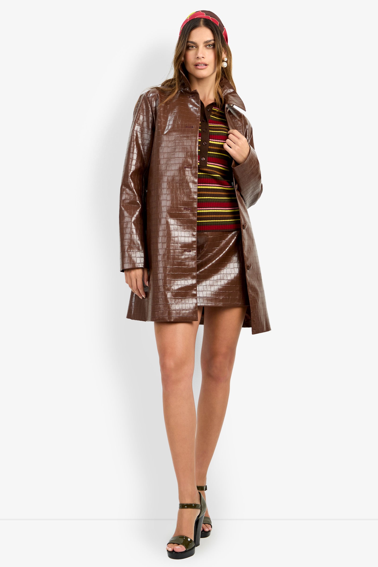 Wearing the DIVIAN COAT, a woman stands against a plain white background, styled with a striped mini dress, black platform sandals, and a red headband.