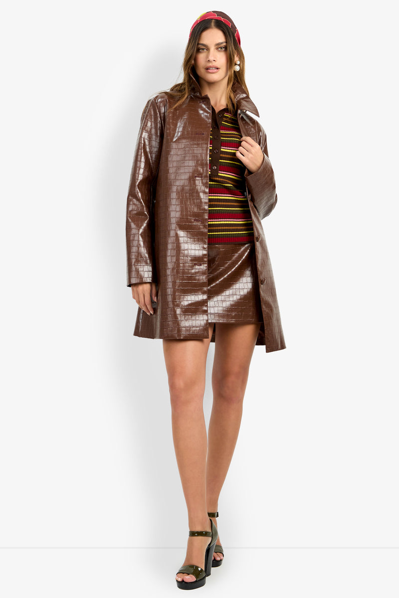 Wearing the DIVIAN COAT, a woman stands against a plain white background, styled with a striped mini dress, black platform sandals, and a red headband.