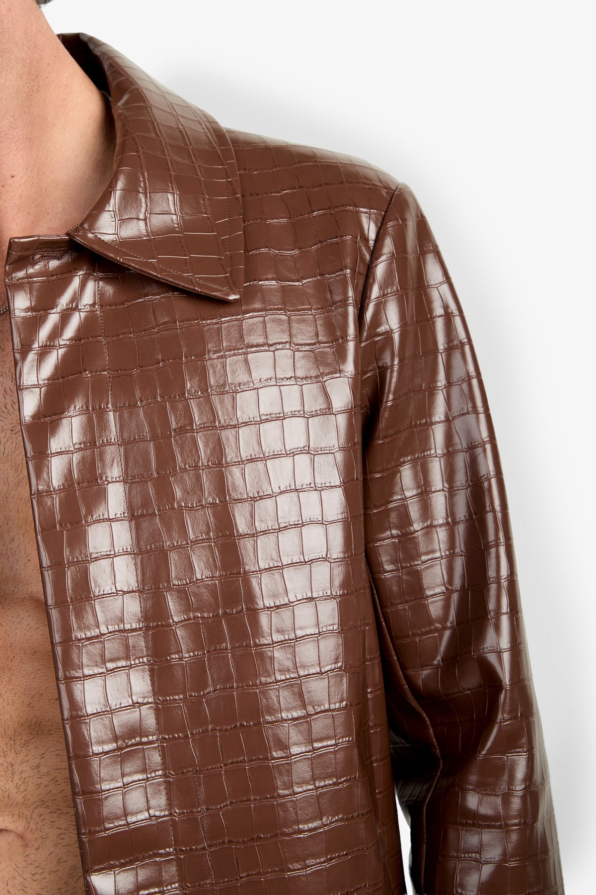 A close-up of a person wearing the Morton Trench Coat, a brown, shiny, crocodile-patterned jacket with an open collar that reveals part of their bare chest, set against a plain white background.