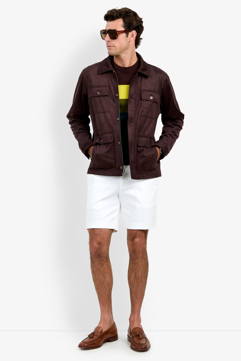 A man wearing sunglasses, the Highland Park Jacket, a dark shirt with a yellow stripe, white shorts, and brown loafers stands with his hands in his pockets against a white background.
