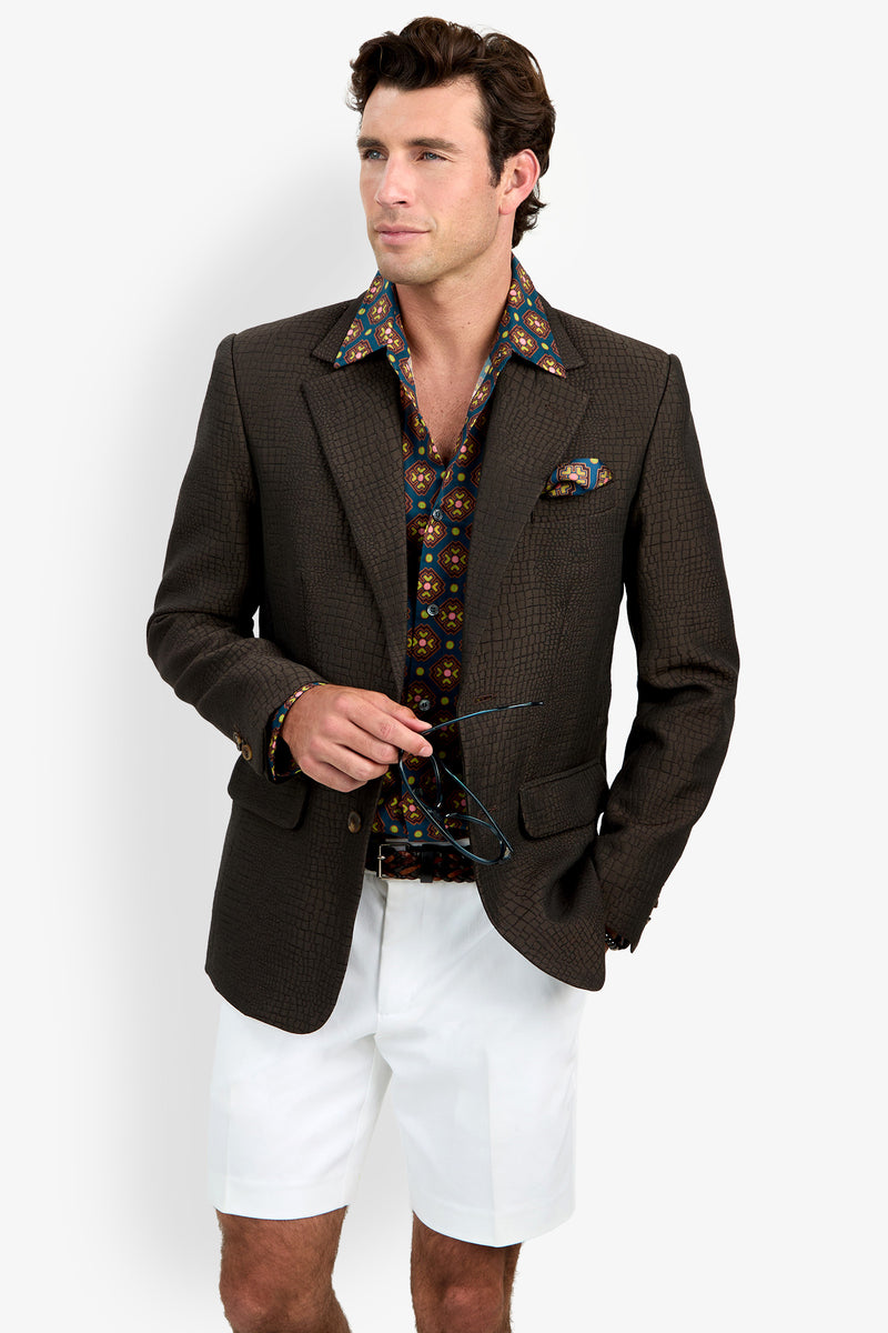 A man wearing the Dohney Blazer, paired with a colorful patterned shirt, a matching pocket square, white shorts, and holding sunglasses, stands against a plain background.