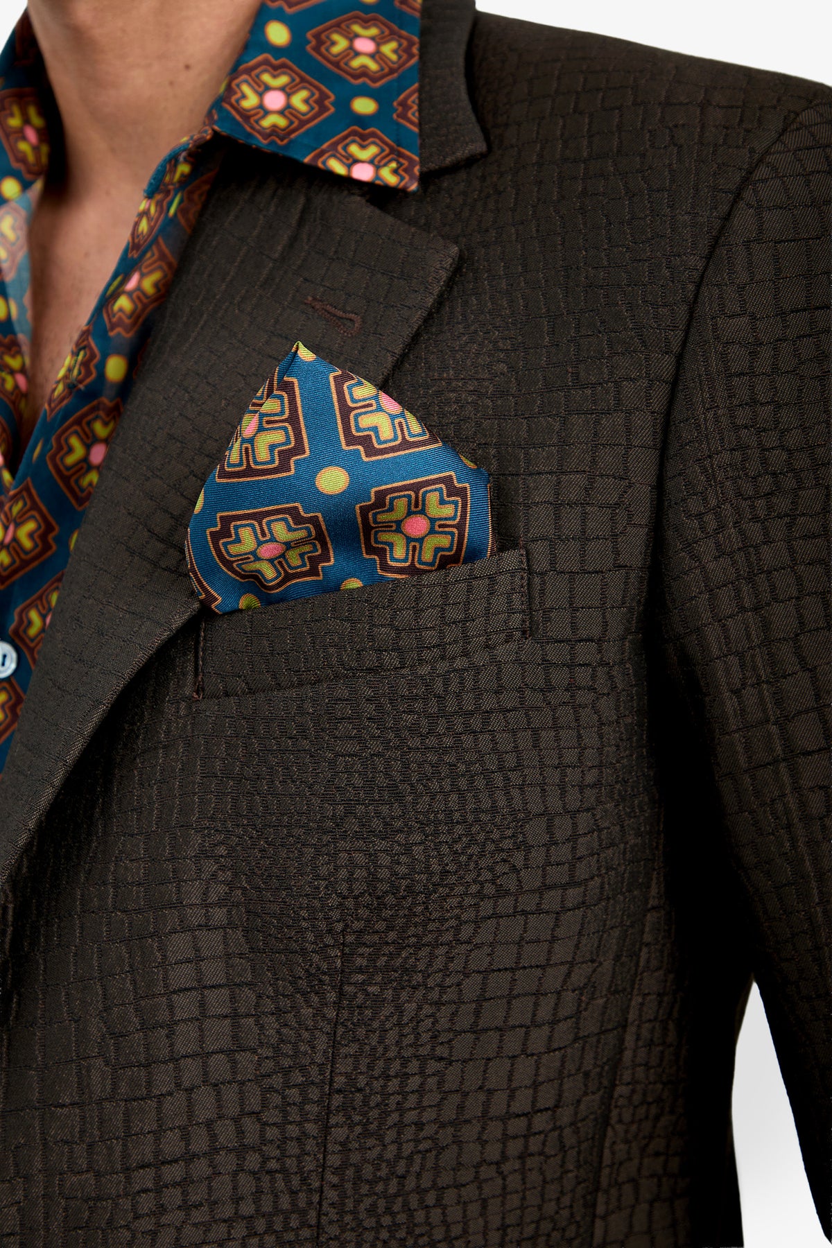 A close-up of a person wearing the Dohney Blazer, featuring a textured dark fabric paired with a colorful geometric-patterned shirt and matching pocket square. The ensemble showcases bold, intricate designs.