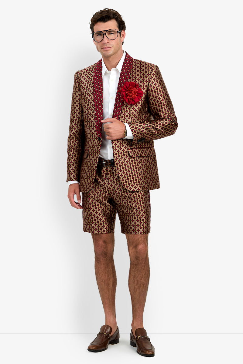 A man wearing glasses, the Alfred Blazer—a patterned brown and red suit jacket with matching shorts—a white shirt, a red flower on the lapel, a brown belt, and brown loafers stands against a plain white background.