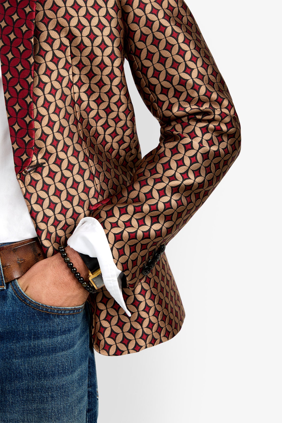 A person wearing blue jeans, a white shirt, and the Alfred Blazer—a patterned red, gold, and black jacket—with one hand in their pocket, displaying a black beaded bracelet.