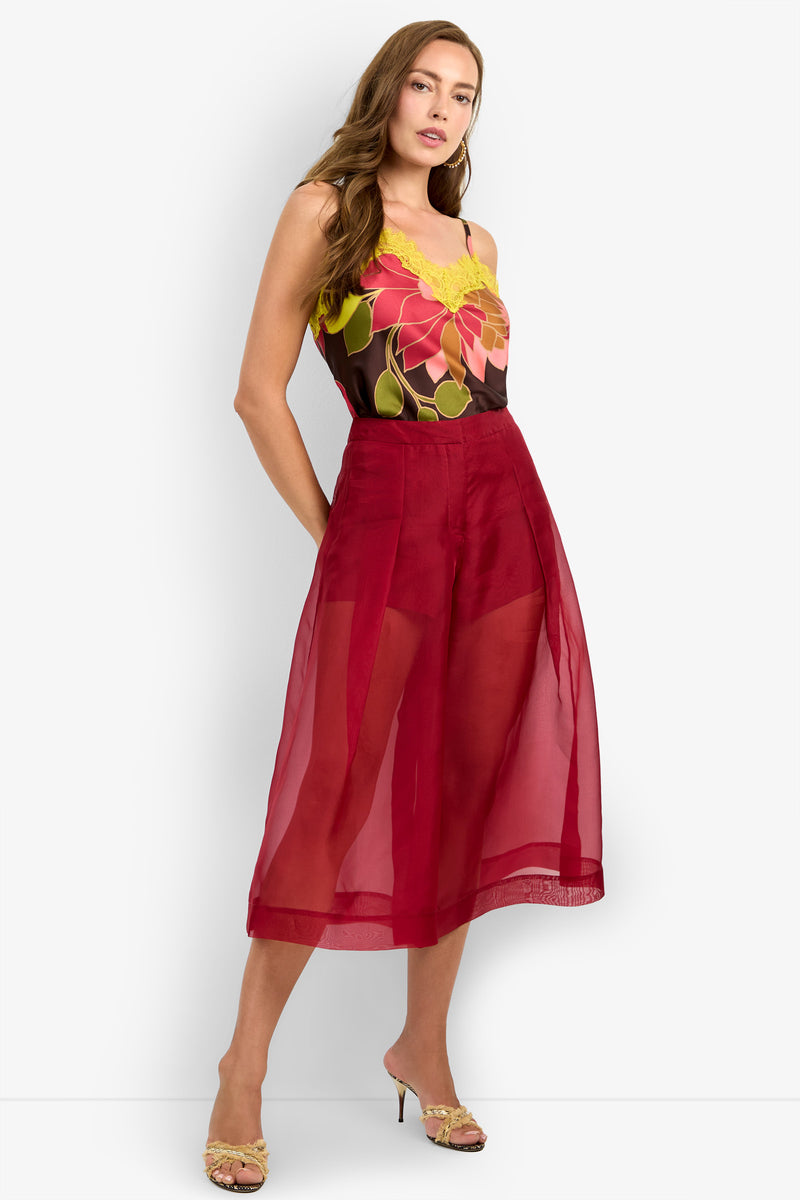 Woman wearing the SIENA PANT, standing with hands in pockets and looking at the camera. She has long brown hair and is styled with a floral sleeveless top and high-heeled sandals.