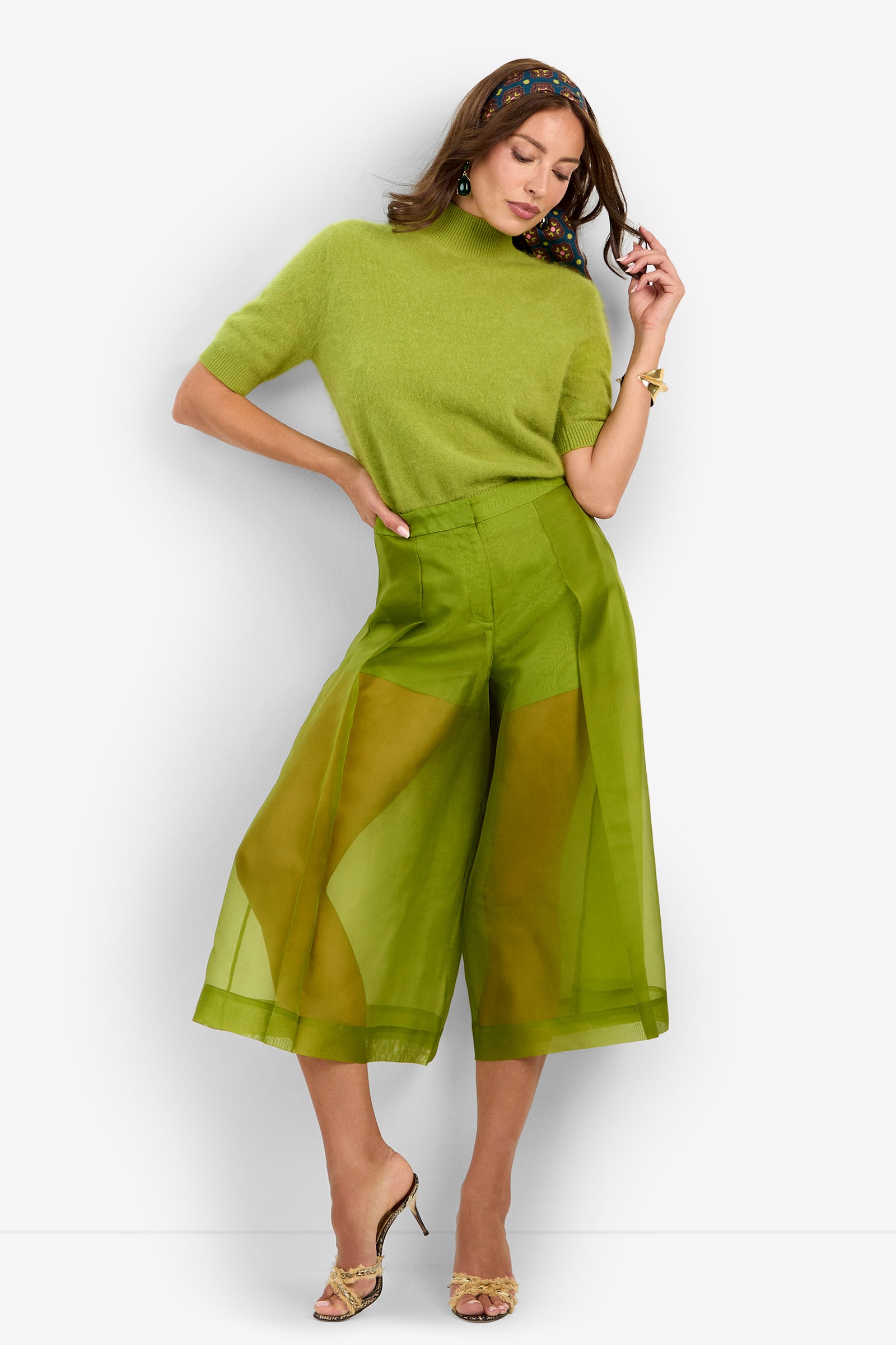 A woman stands against a white background in heels, wearing a light green short-sleeve sweater and the Siena Pant—sheer, wide-leg green trousers—with one hand on her head and the other at her waist.