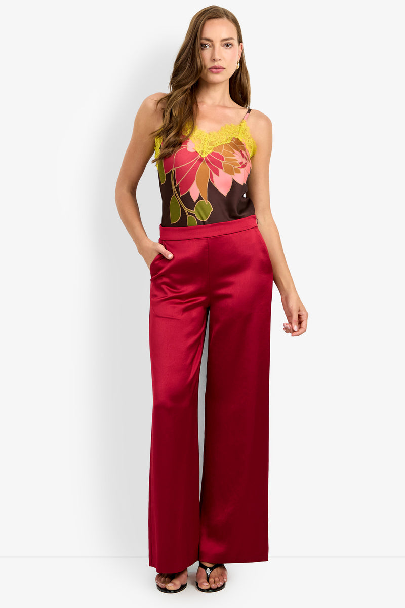A woman stands against a plain background with one hand in the pocket of her INES PANT, wide-leg and shiny red, paired with a sleeveless floral top featuring yellow lace accents.