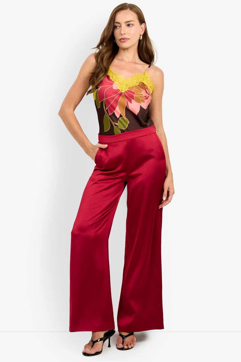 A woman wearing the INES PANT in red satin, paired with a sleeveless floral top featuring yellow lace trim and black heels, stands with one hand in her pocket against a white background.