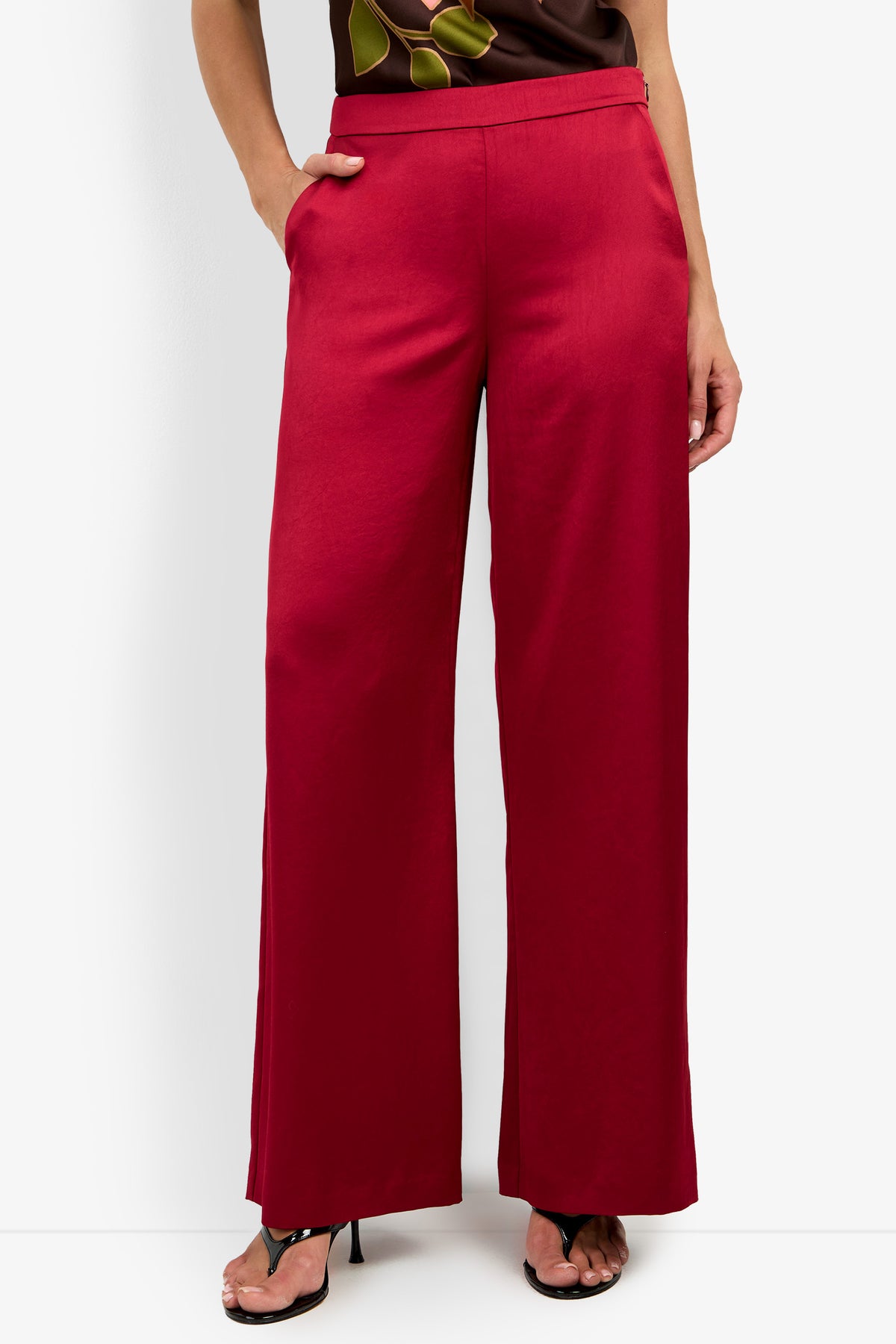A person models the INES PANT, a pair of high-waisted, wide-leg red pants, paired with black open-toe heels against a white background. Only the lower half of their body is shown.