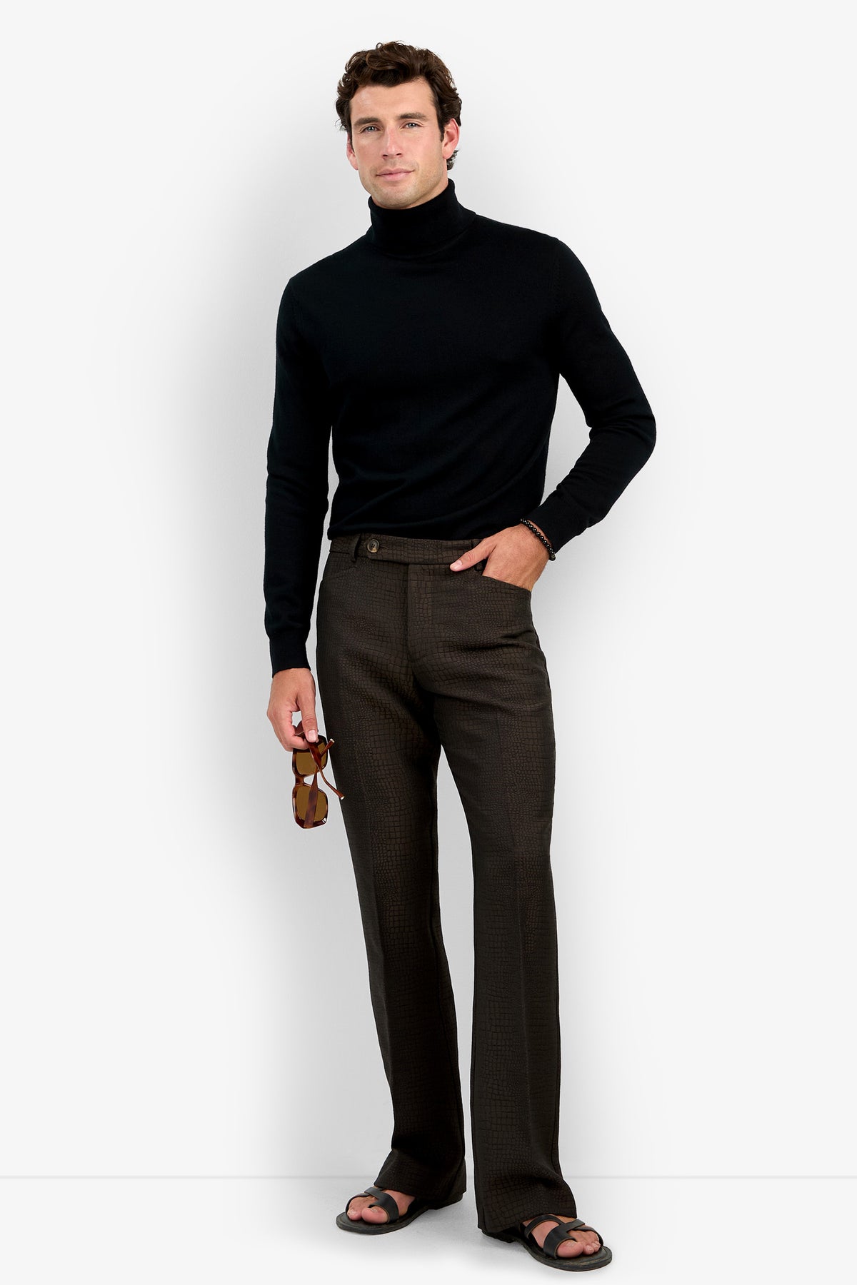 A man wearing a black turtleneck sweater, David Trouser in brown plaid, and black sandals holds sunglasses in his right hand while standing against a plain white background.
