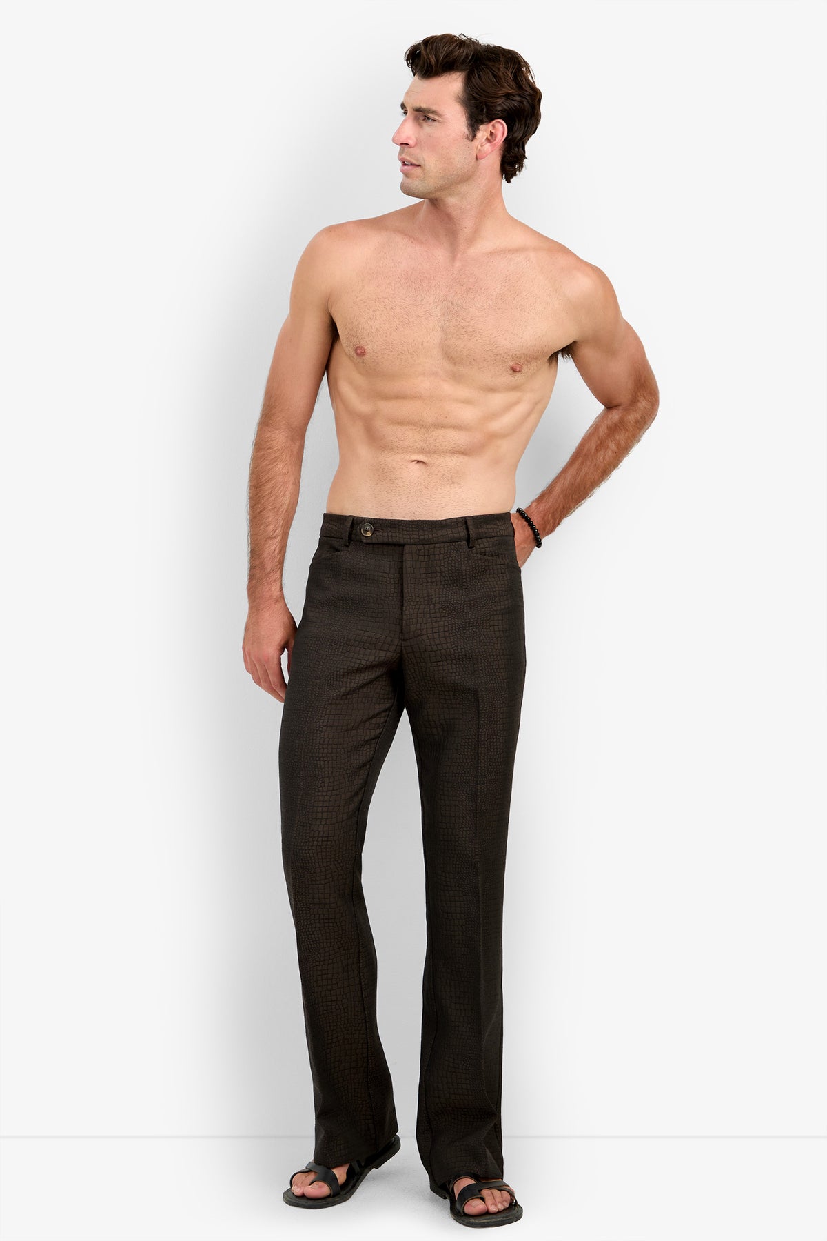 A shirtless man wearing the David Trouser and black sandals stands against a plain white background, looking to his left with one hand resting on his hip.