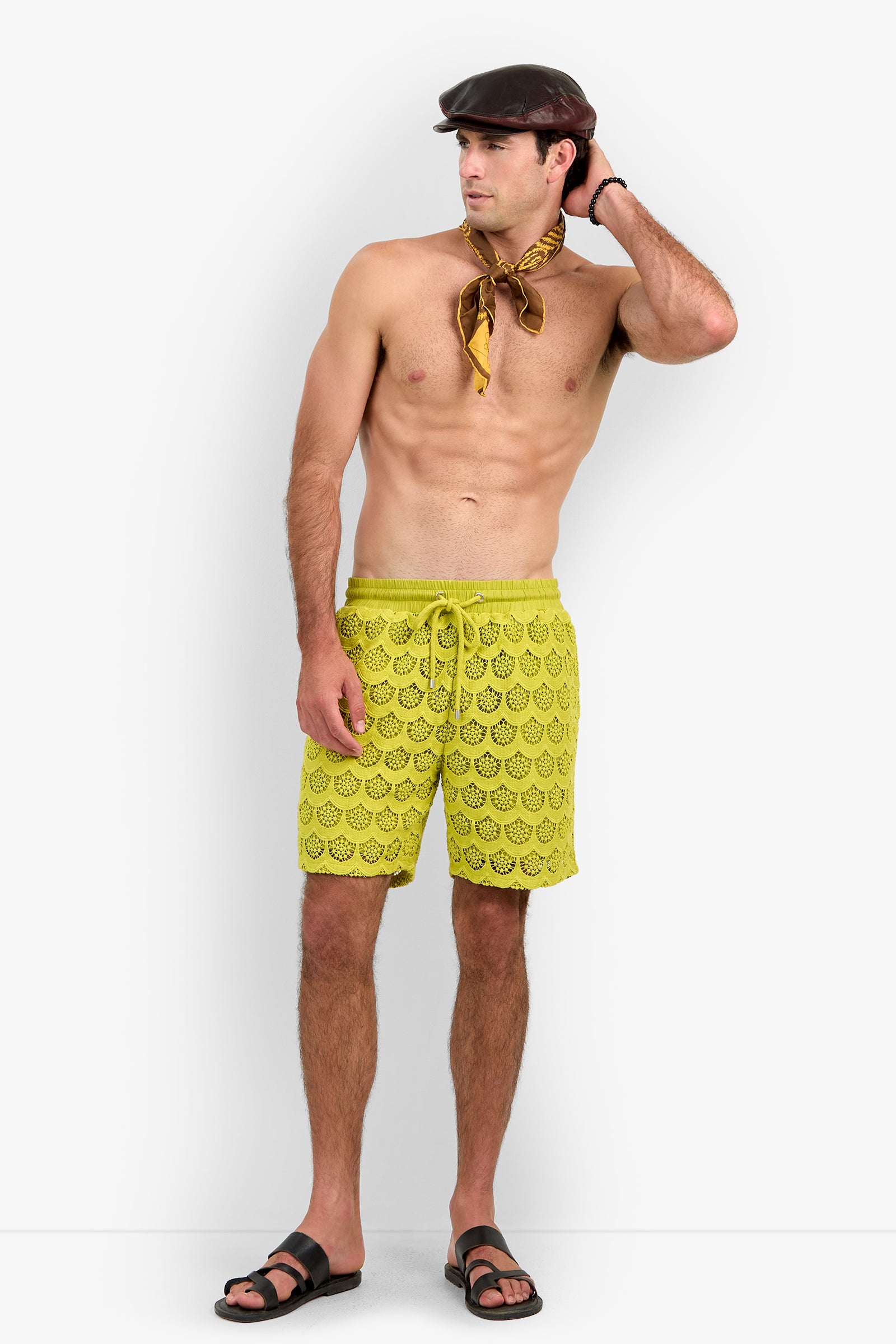 A shirtless man wearing the Shoreline Short in yellow, paired with black sandals, a brown flat cap, and a yellow scarf around his neck, stands with one hand on his head looking to the side against a plain white background.