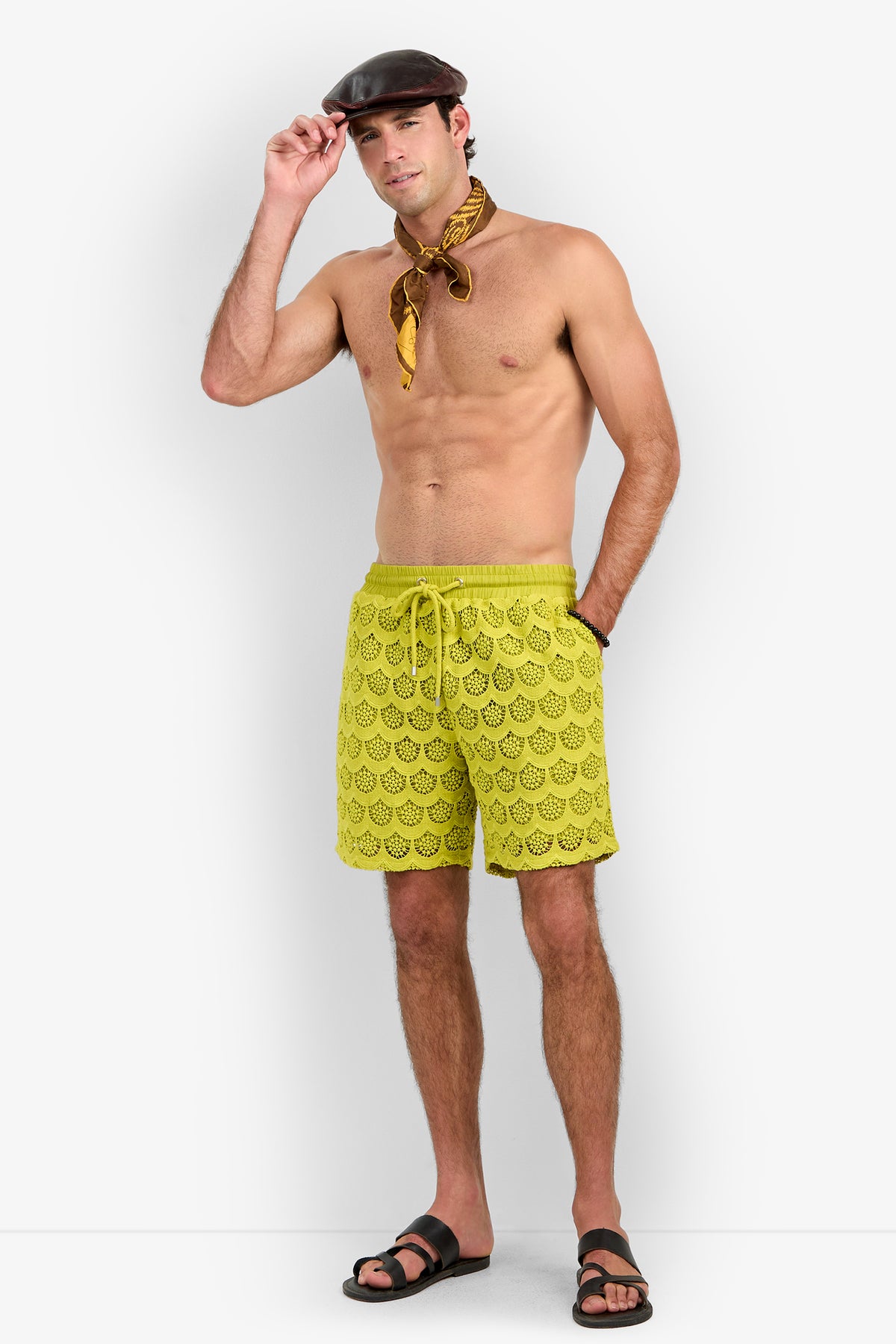 A man wearing a brown cap, yellow scarf, Shoreline Short swim trunks with a yellow pattern, and black sandals stands against a white background with one hand touching his cap and the other in his pocket.