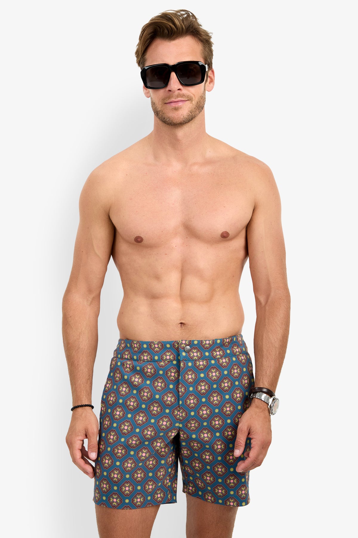 A man wearing the HONOLULU BOARDSHORT, featuring a stylish pattern, stands shirtless against a plain white background. He has short hair and accessorizes with sunglasses, a watch, and a bracelet on his left wrist.