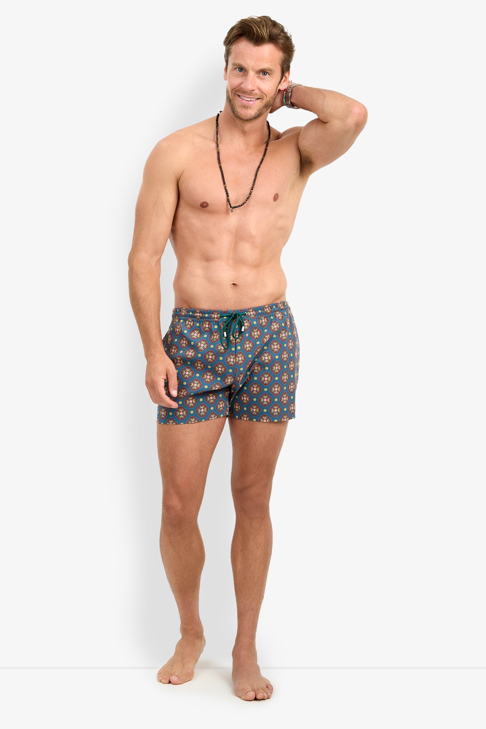 A smiling man with a short beard stands barefoot in the OCEANSIDE SWIM TRUNK, wearing a black necklace, with one hand touching the back of his head against a plain white background.
